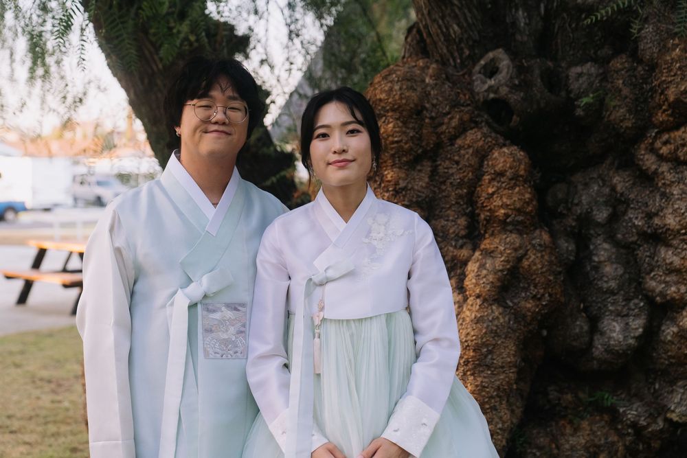 A couple wearing traditional Korean hanbok attire poses together beside a large tree outdoors.