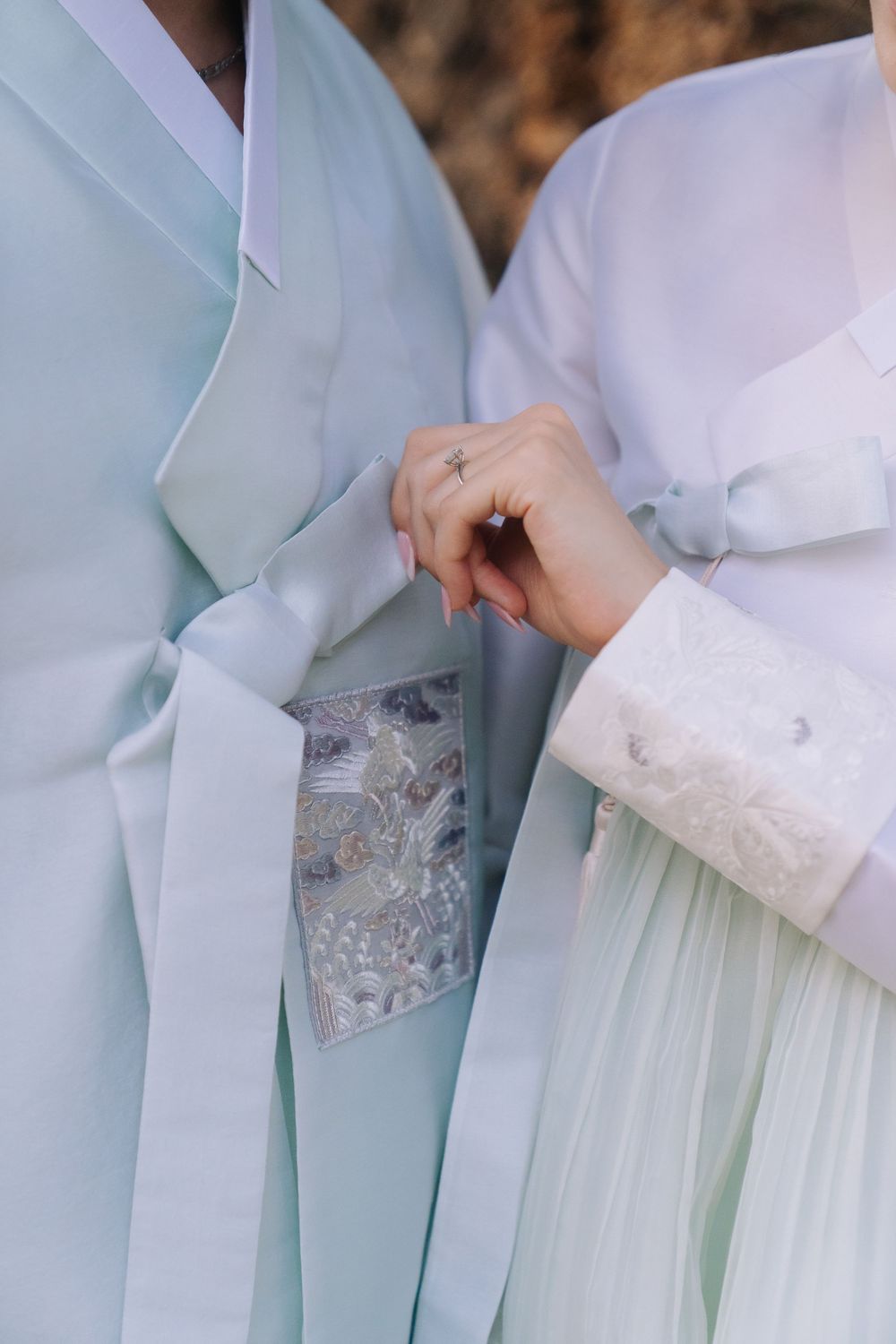 Close-up of couple holding hands in pastel Korean hanbok with intricate embroidered details at wedding.