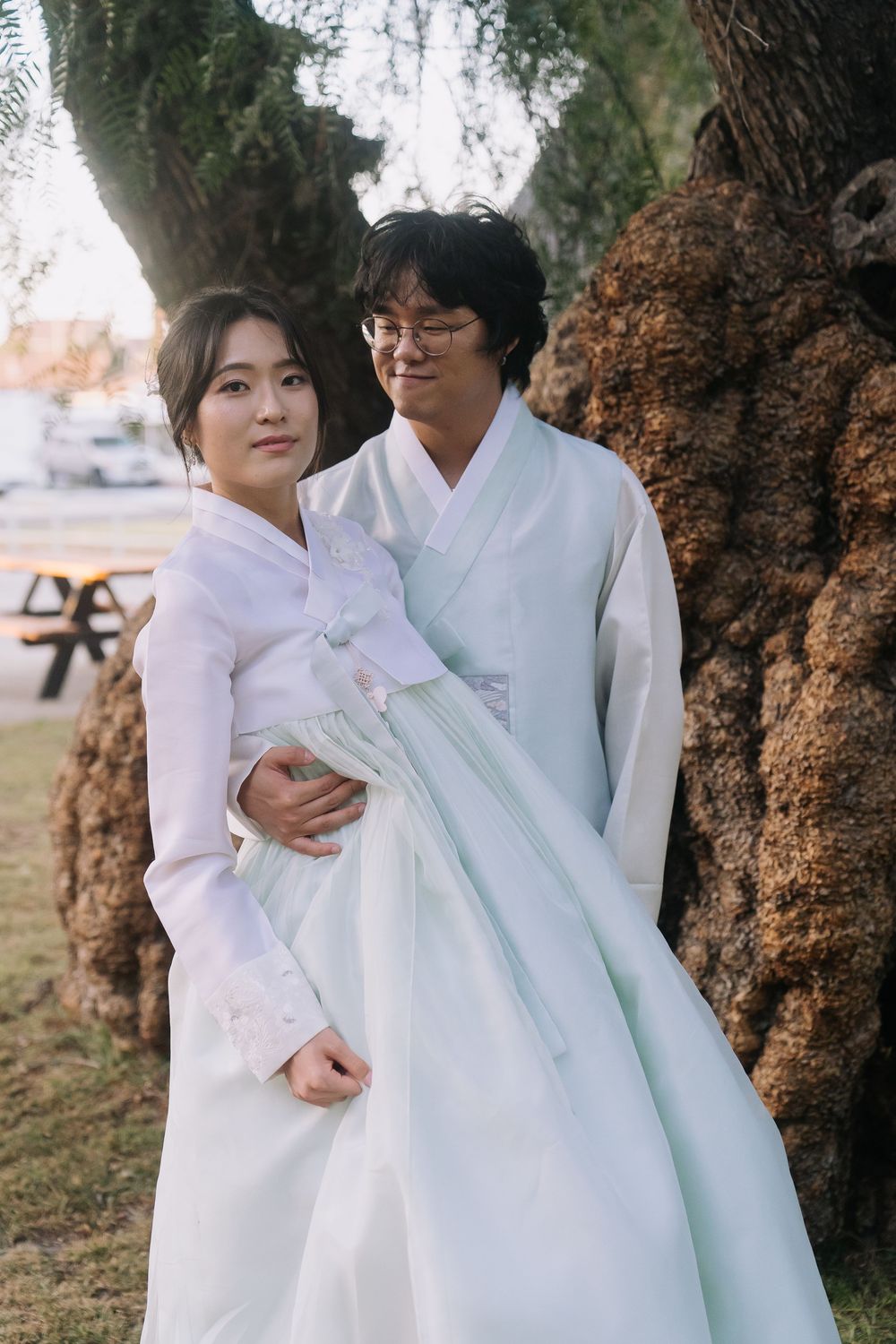 Korean couple in matching white and mint hanbok posing together beside an ancient tree outdoors.