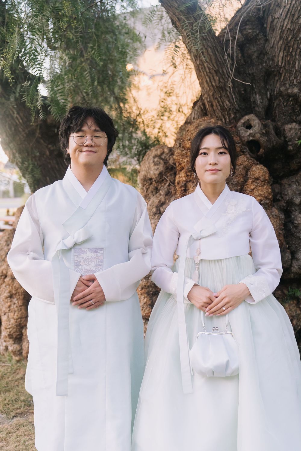 Couple wearing traditional white Korean hanbok standing together outdoors near a large tree.