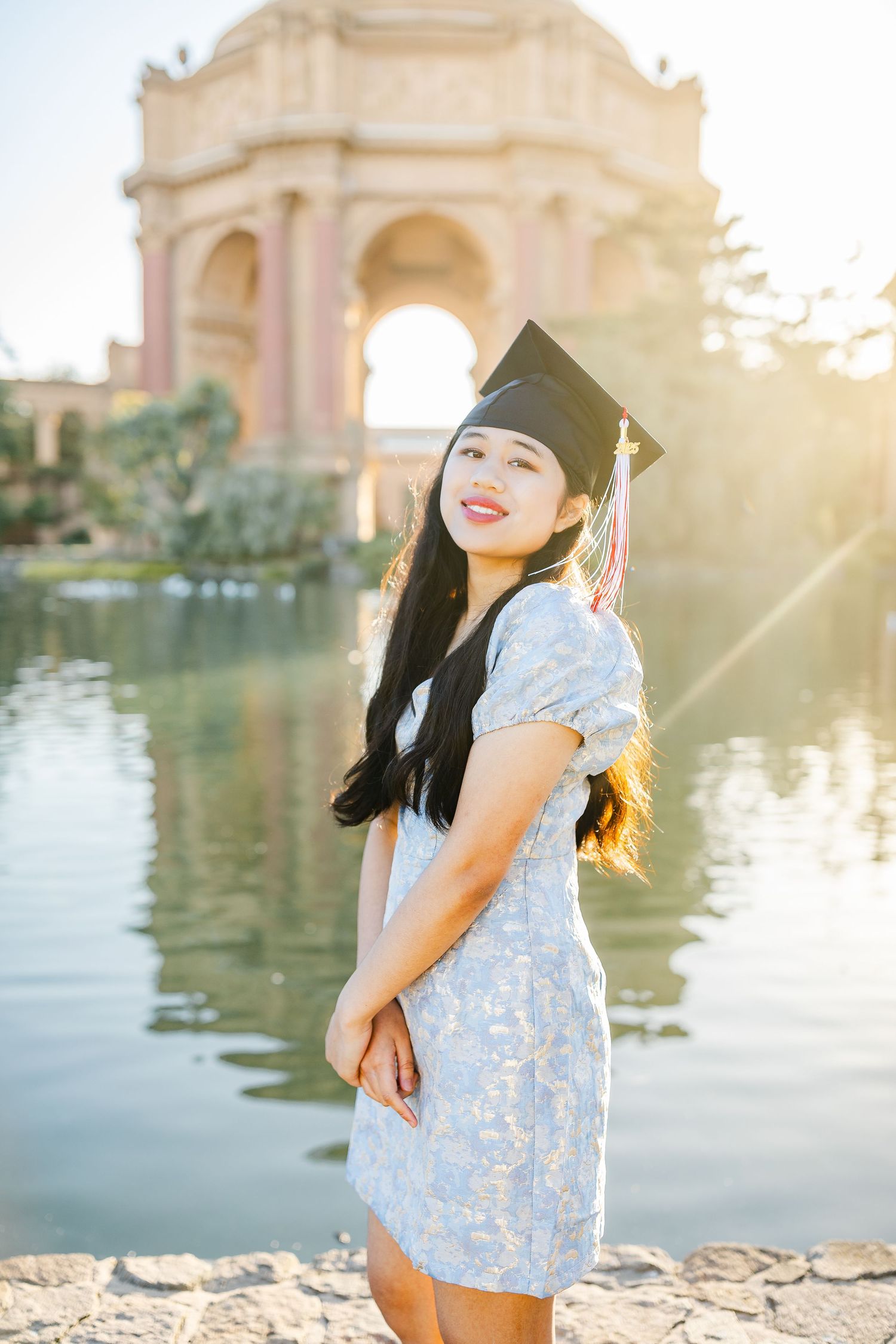 Young woman in graduation cap and floral dress smiling by a pond with classical arch architecture in background.