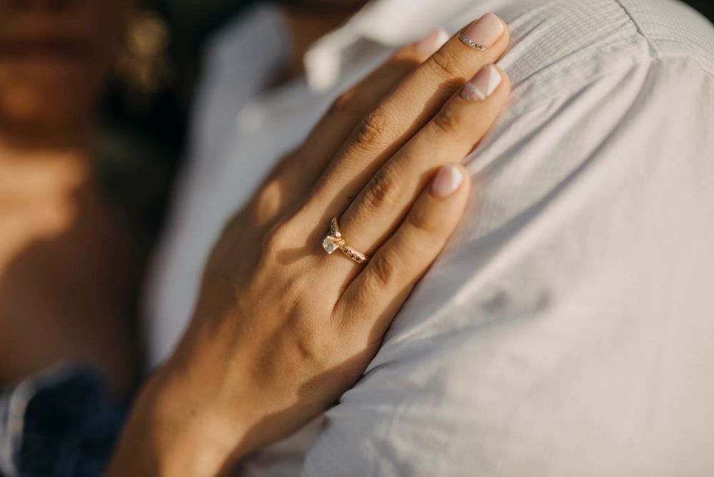 Close-up of a woman's hand wearing a solitaire diamond engagement ring resting on a man's shoulder.