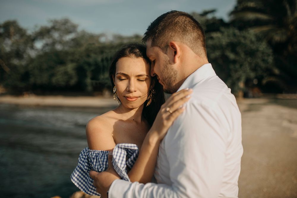 Romantic couple embracing on a beach, woman in off-shoulder top, man in white shirt, with tropical trees in background.