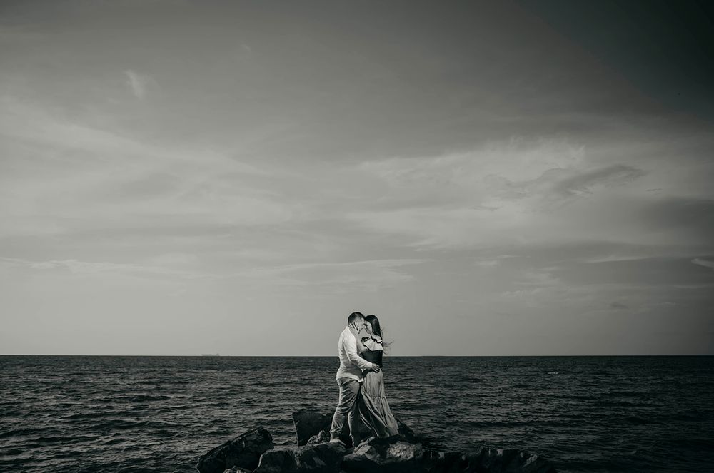 Black and white photo of a couple embracing on rocks by the ocean under a dramatic cloudy sky.