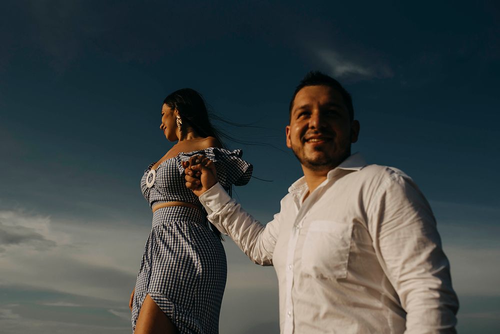 A couple holds hands under a dramatic blue sky, woman in striped dress with wind-blown hair, man in white shirt smiling.