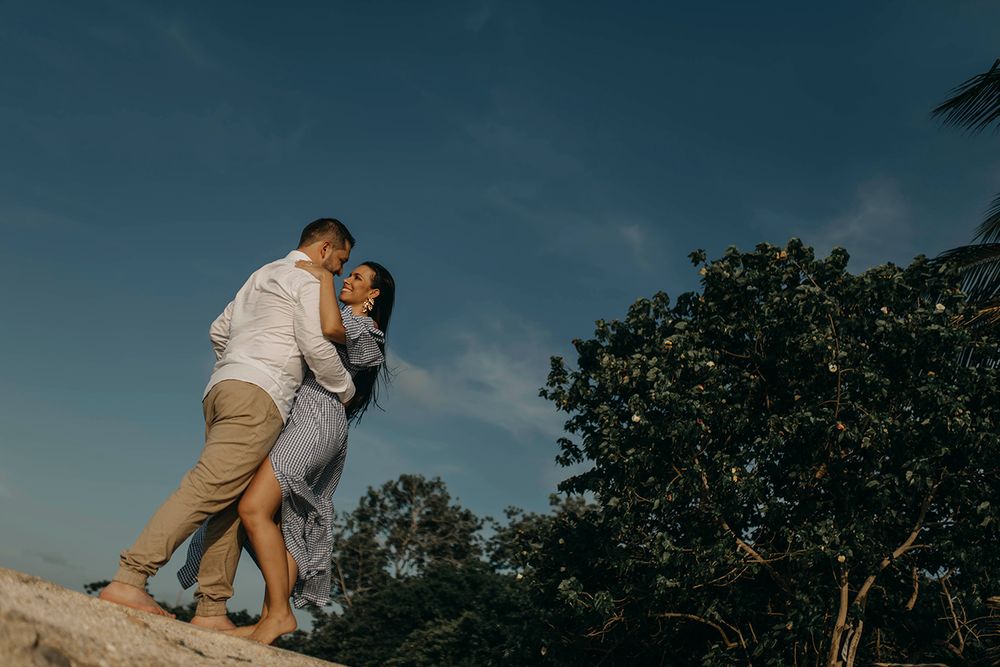 Couple embracing romantically on rocks outdoors under a blue sky with lush green trees in the background.