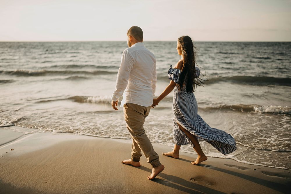 Couple holding hands walking barefoot on sandy beach at sunset, waves gently rolling in behind them.