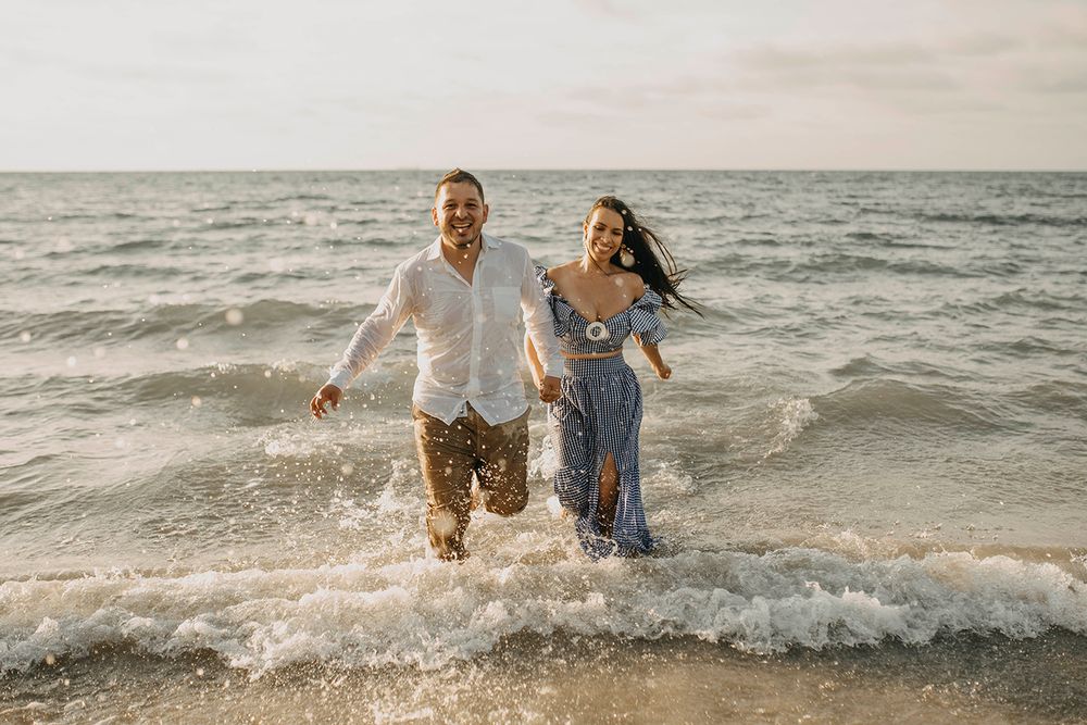 Couple laughing and running through ocean waves on a sunny beach, man in white shirt and woman in blue dress.