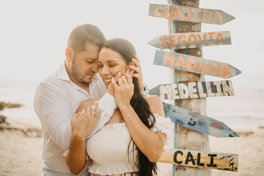 Couple sharing a tender moment beside a colorful wooden directional sign on a sunny beach.