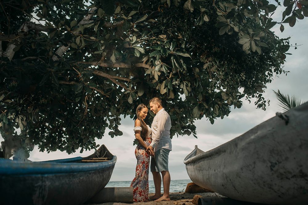 Romantic couple embracing under a large tropical tree on a beach with wooden boats nearby.