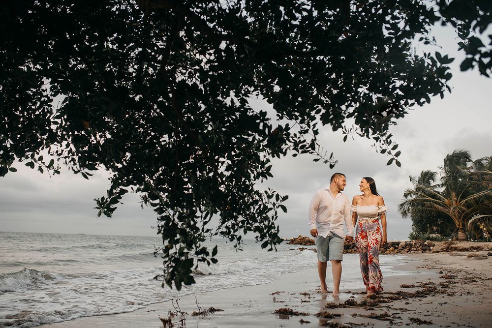 Couple walking hand in hand on a tropical beach shore under lush overhanging trees with ocean waves in background.