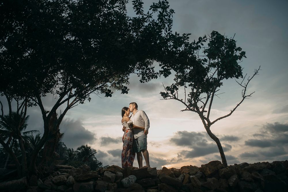 Couple embracing at sunset under trees on rocky terrain with dramatic cloudy sky backdrop.