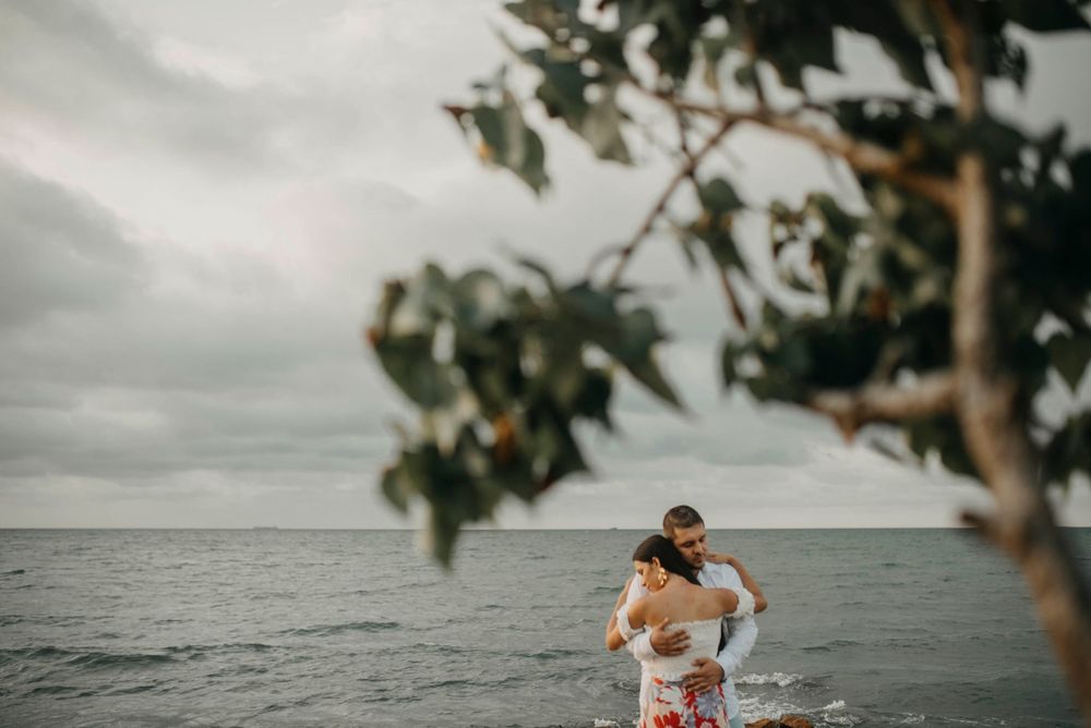Couple embracing romantically by the ocean shore with lush green tree branches framing the scene under a cloudy sky.