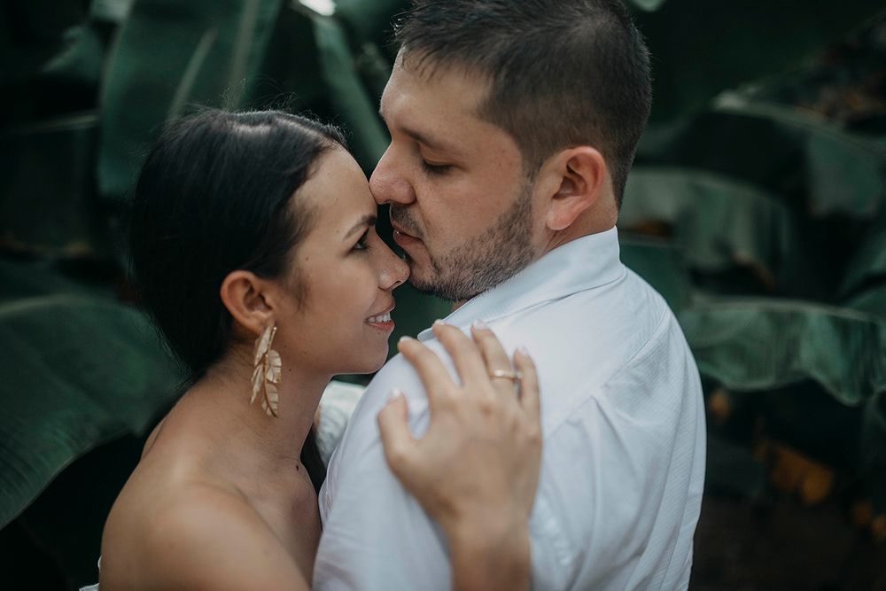 A couple shares a romantic moment, the groom kissing the bride's forehead surrounded by lush tropical greenery.