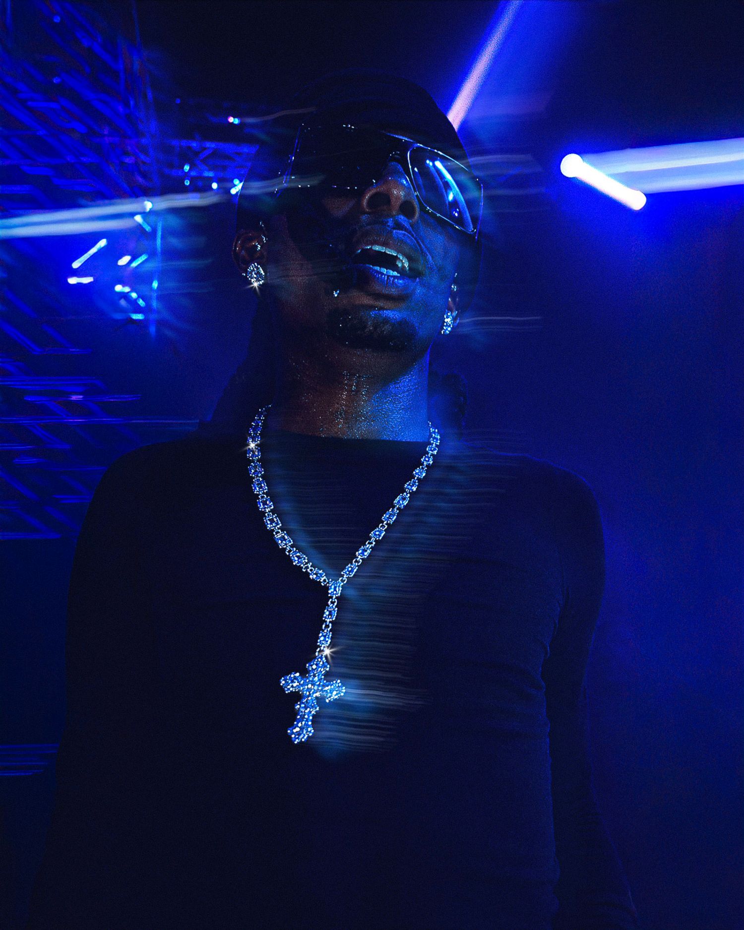 Rapper performing on stage in blue light, wearing sunglasses, cross necklace, and black gloves while holding a microphone.