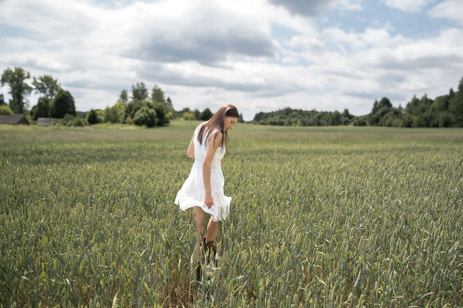 A young woman in a white dress walks through a lush green field with trees in the background.