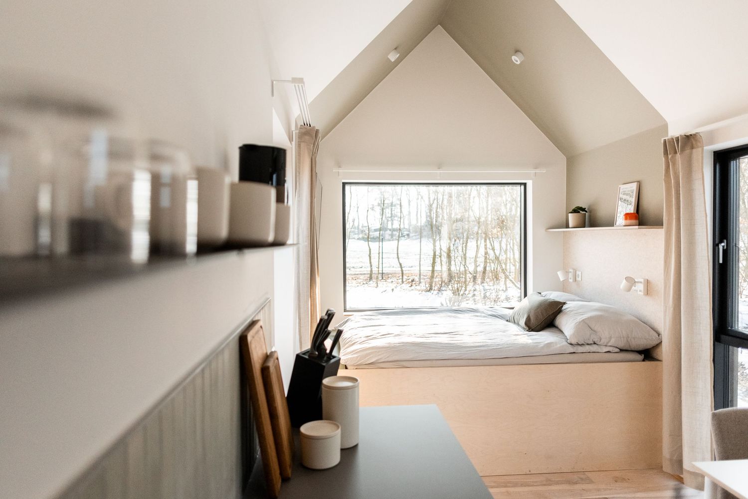 Minimalist tiny home interior with platform bed, large window, vaulted ceiling, and kitchen shelving in neutral tones.