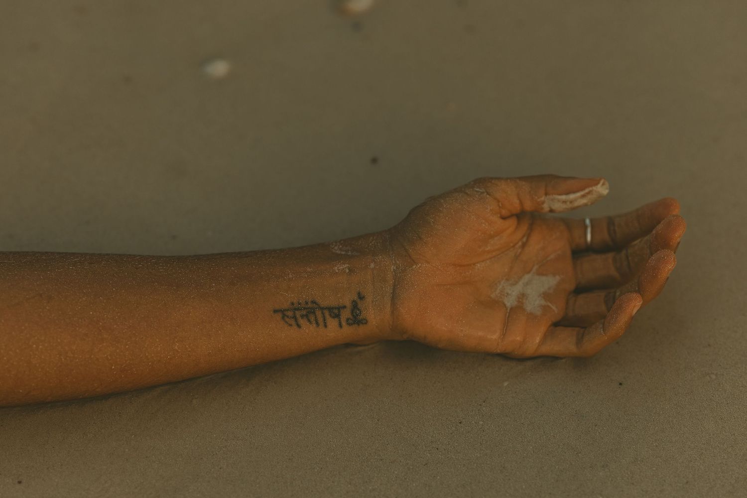 Close-up of a hand with a Sanskrit tattoo on the wrist resting on wet sand at a beach.