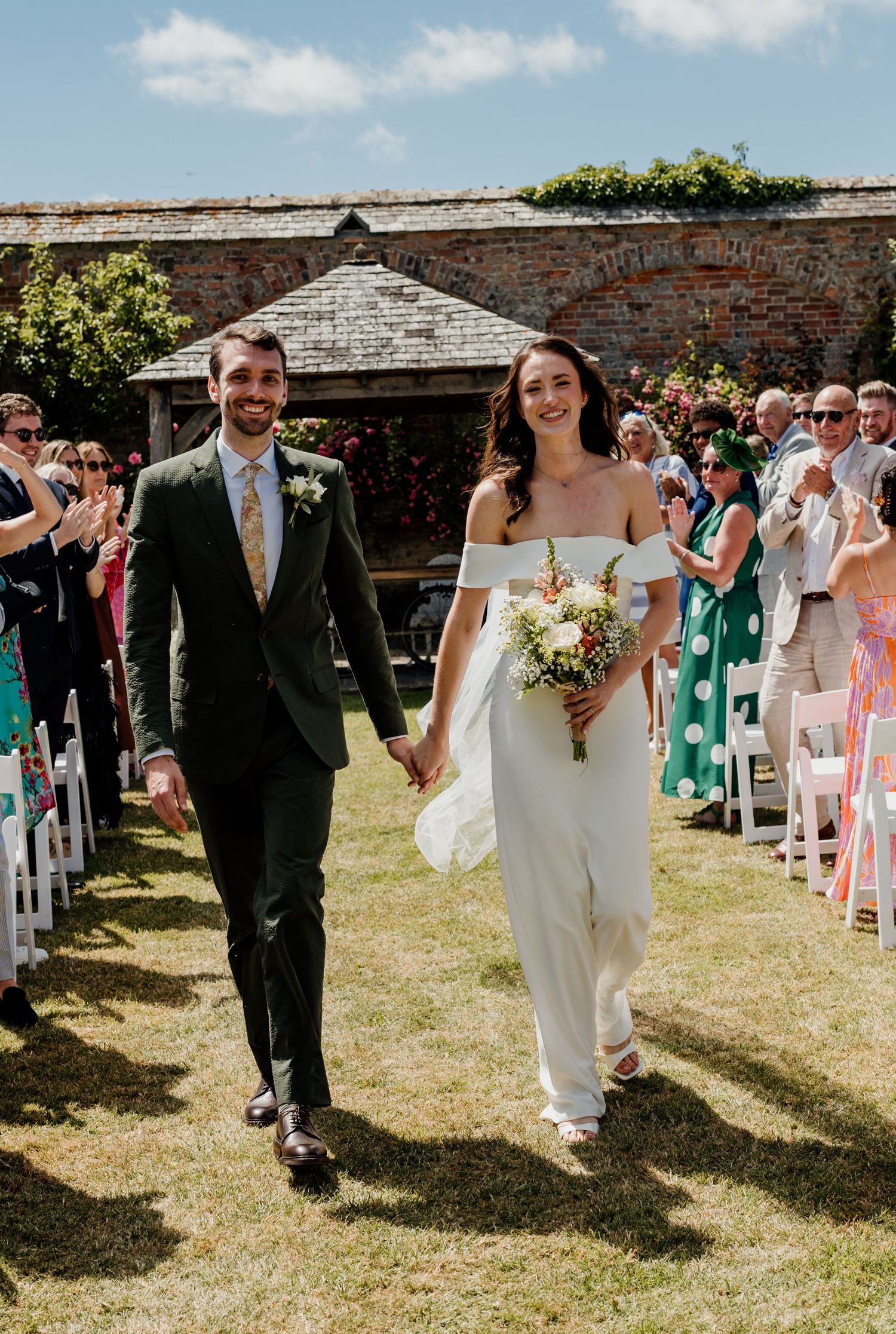 Bride and groom walk hand-in-hand down outdoor aisle after wedding ceremony, guests applauding on sunny day.