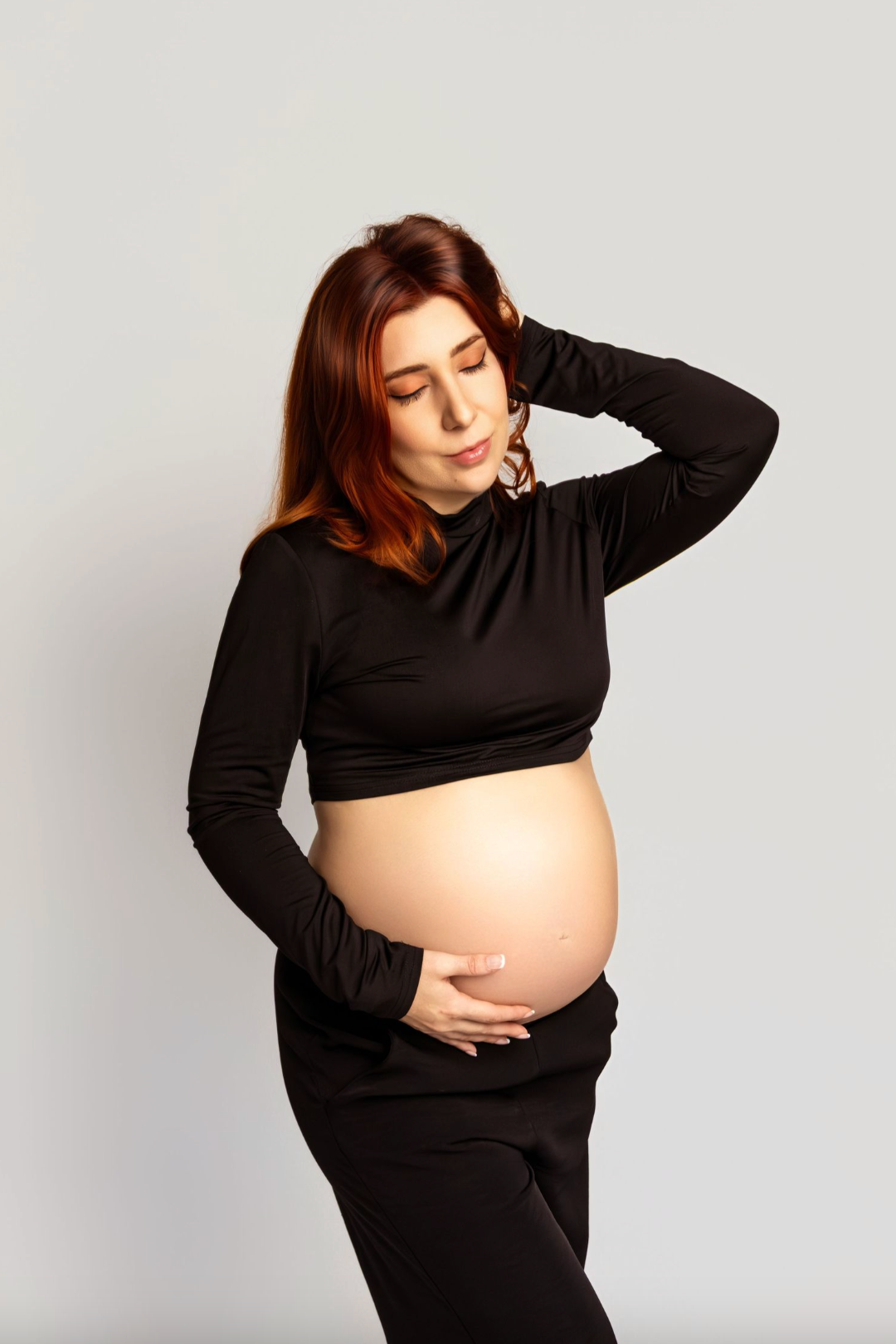 Pregnant woman in black crop top cradling her bare baby bump, posing against a light gray background.
