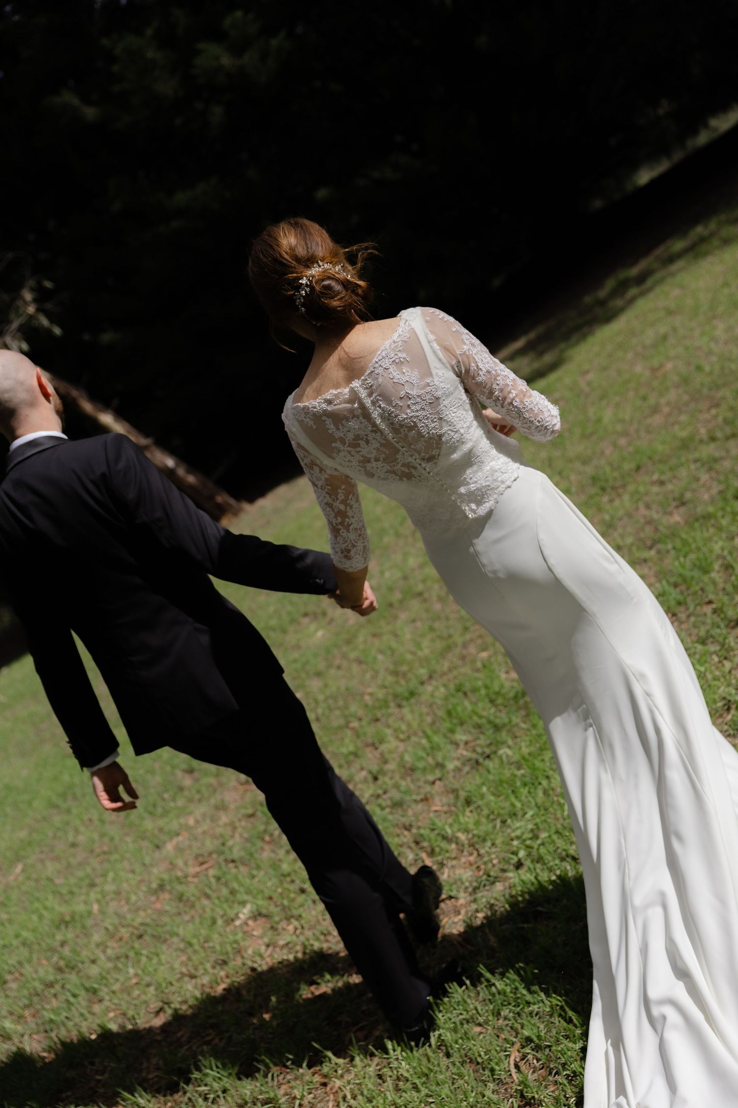 Bride in lace back wedding gown holds groom's hand walking uphill on green grass outdoors.