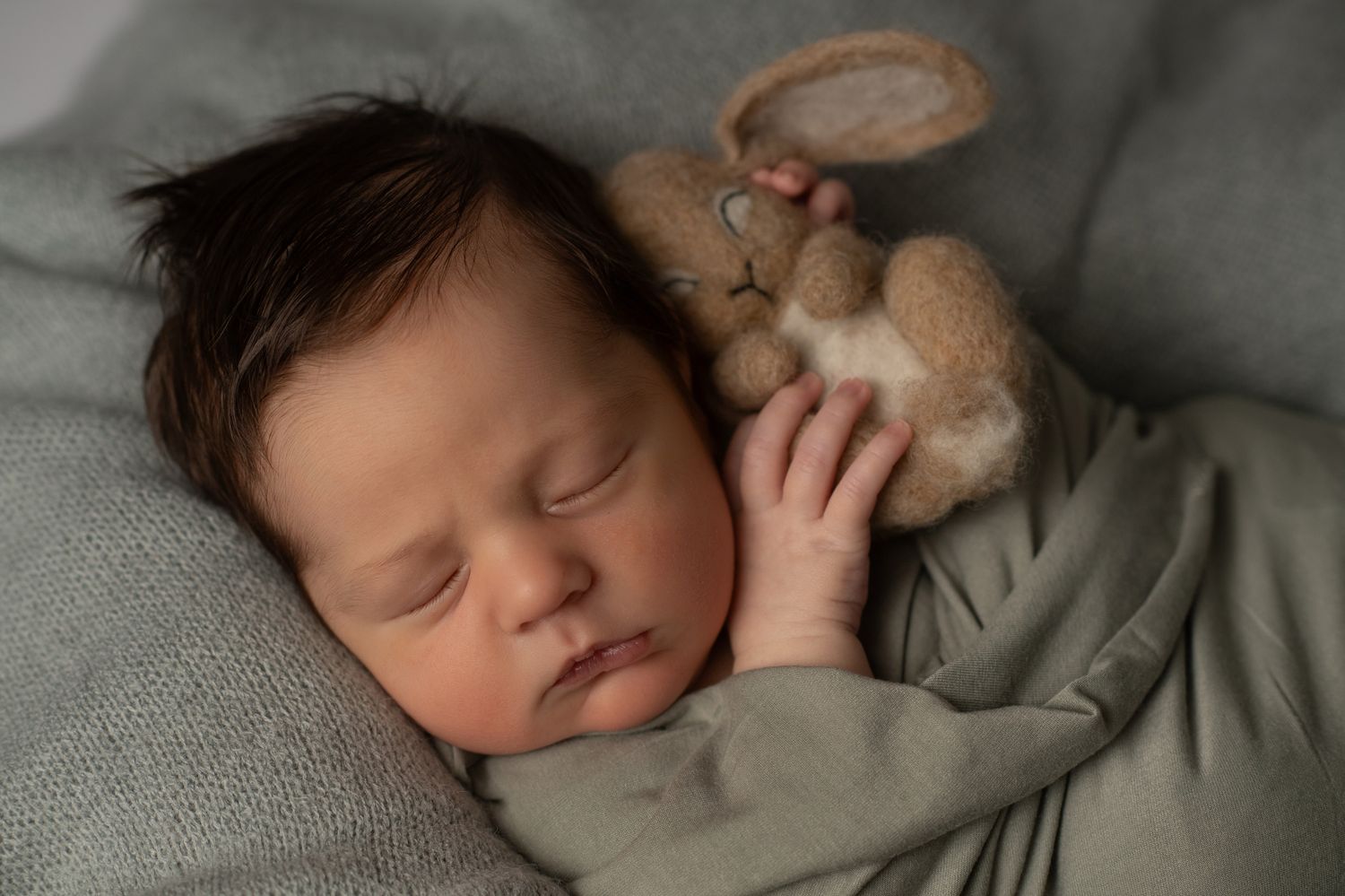 Peaceful newborn baby sleeping with a soft stuffed bunny toy, wrapped in a gray blanket.