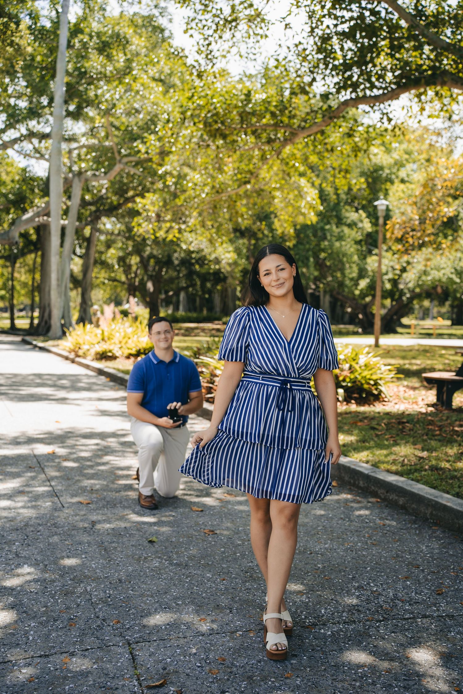 Woman in blue striped dress walks park path while man kneels behind her in sunny green park setting.