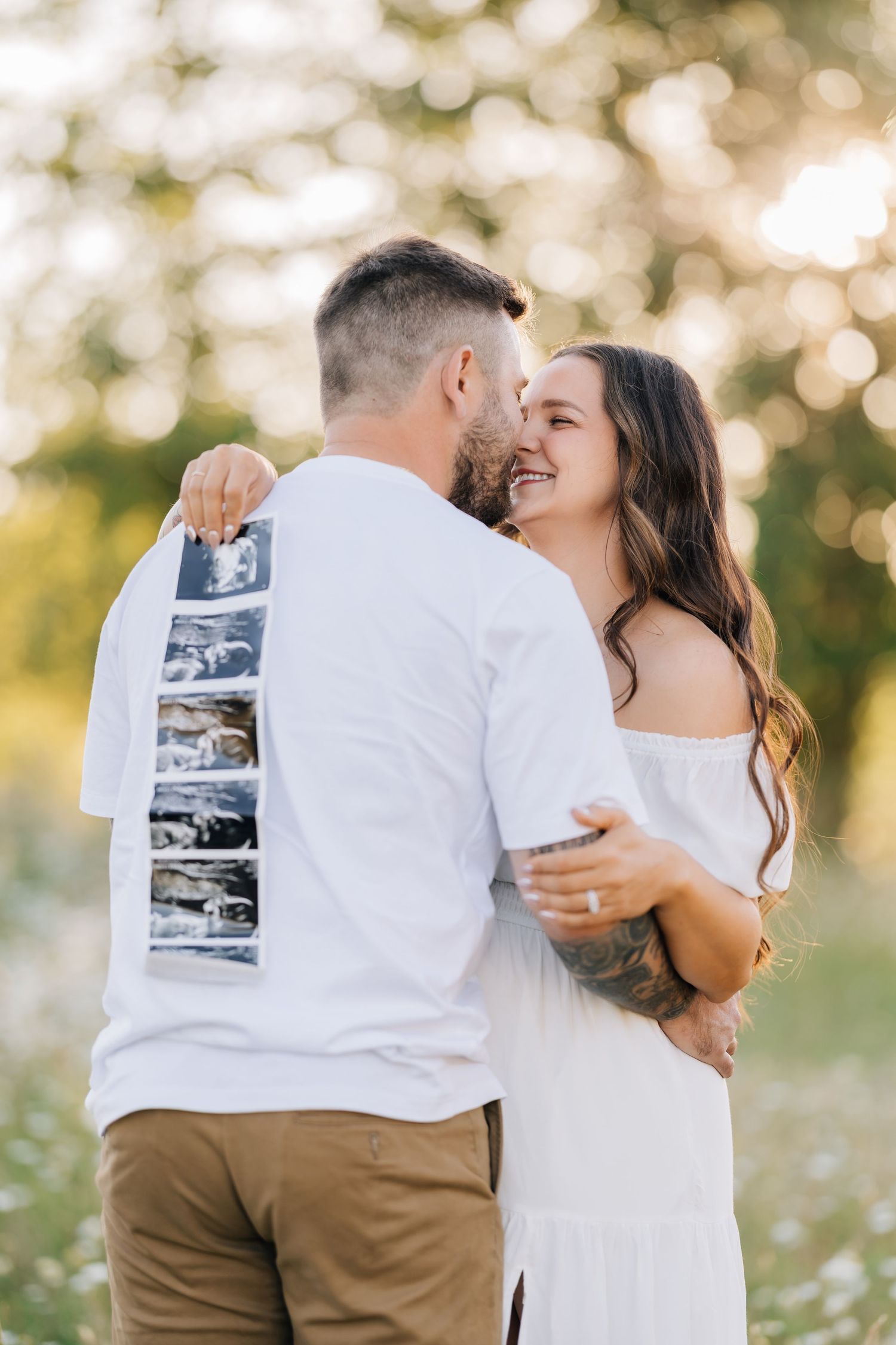 Happy couple embracing outdoors holding ultrasound photos, announcing pregnancy in a sunny field with bokeh background.