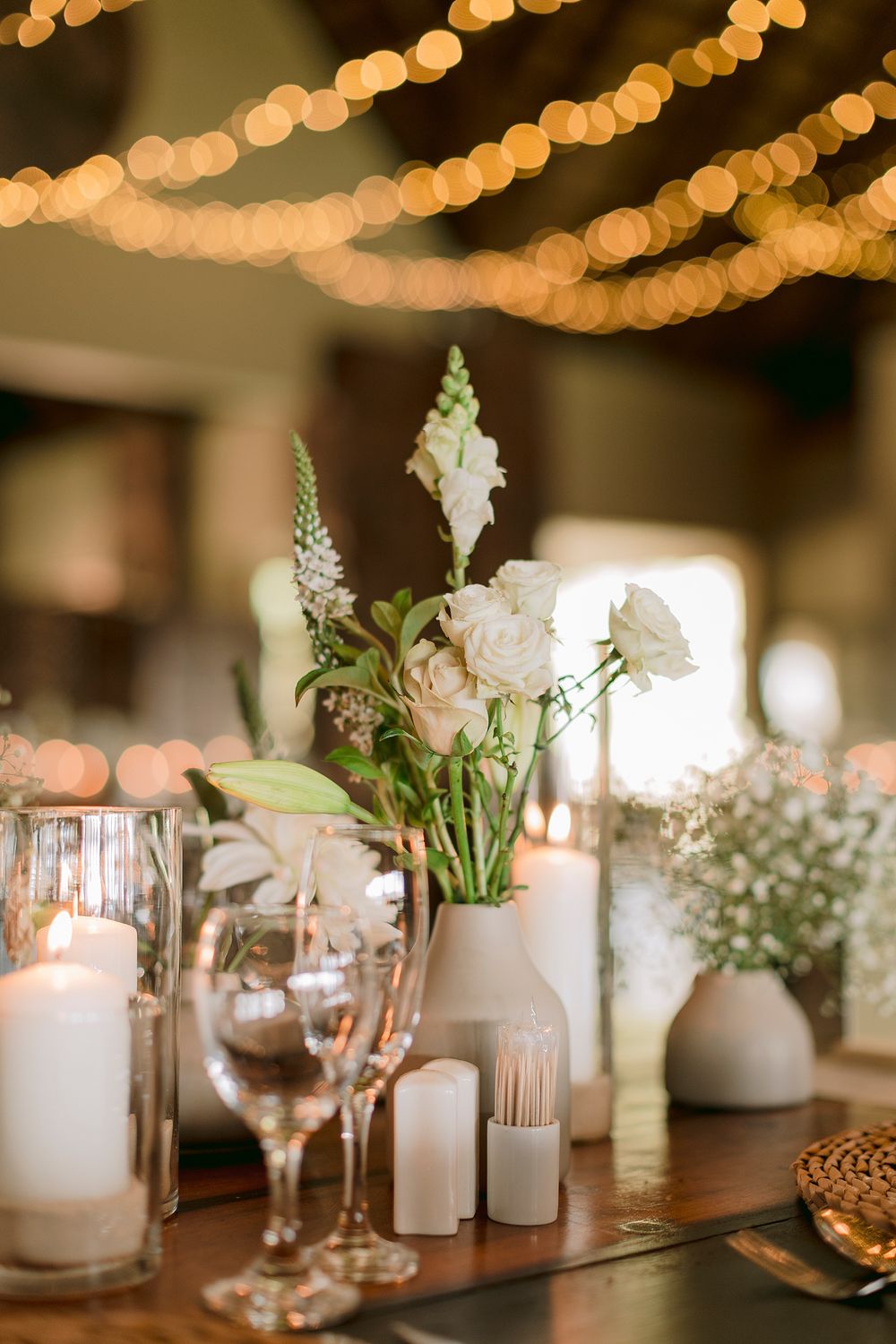 Elegant wedding reception tablescape with white floral centerpieces, candles, and string lights overhead.