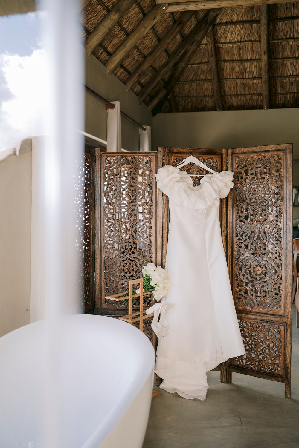White wedding dress with ruffle sleeves hanging on ornate wooden screen near freestanding bathtub in tropical villa.