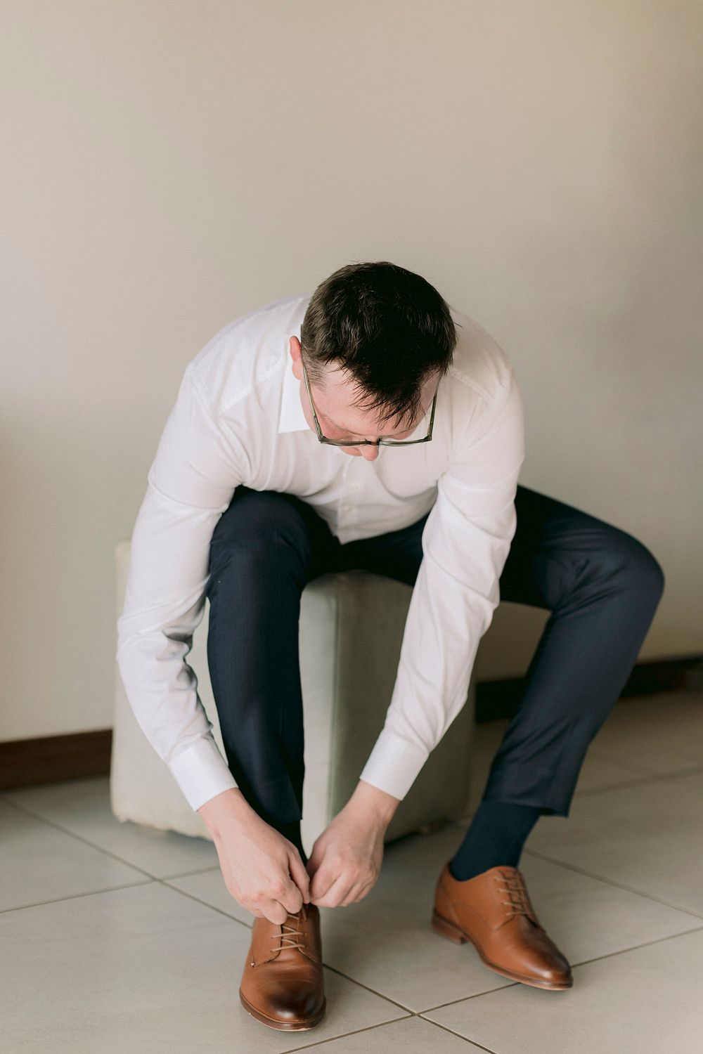 Man in white shirt and navy trousers tying brown oxford shoes while seated, preparing for formal occasion.