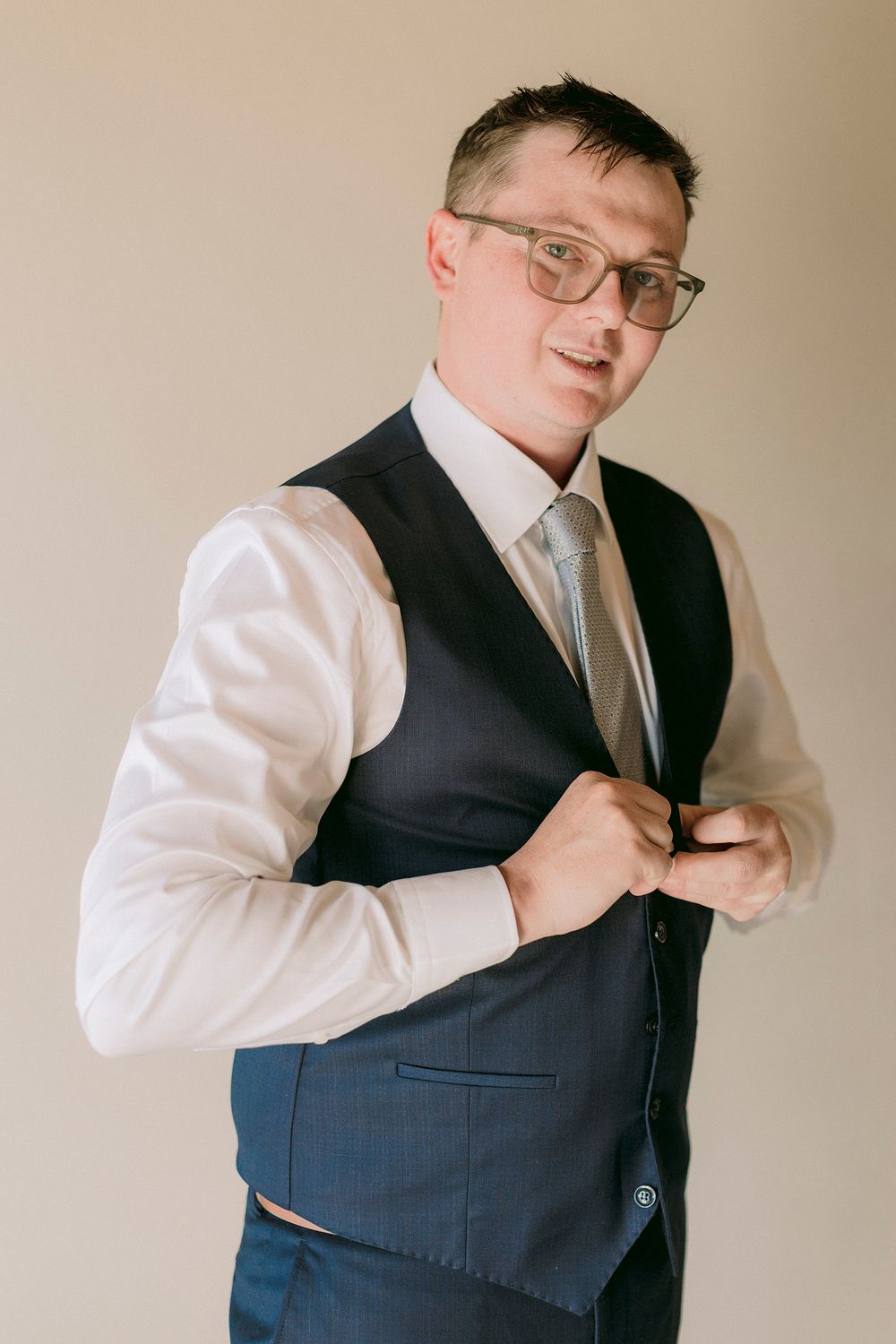 Man in navy vest, white shirt, and tie adjusting his jacket button while wearing glasses.