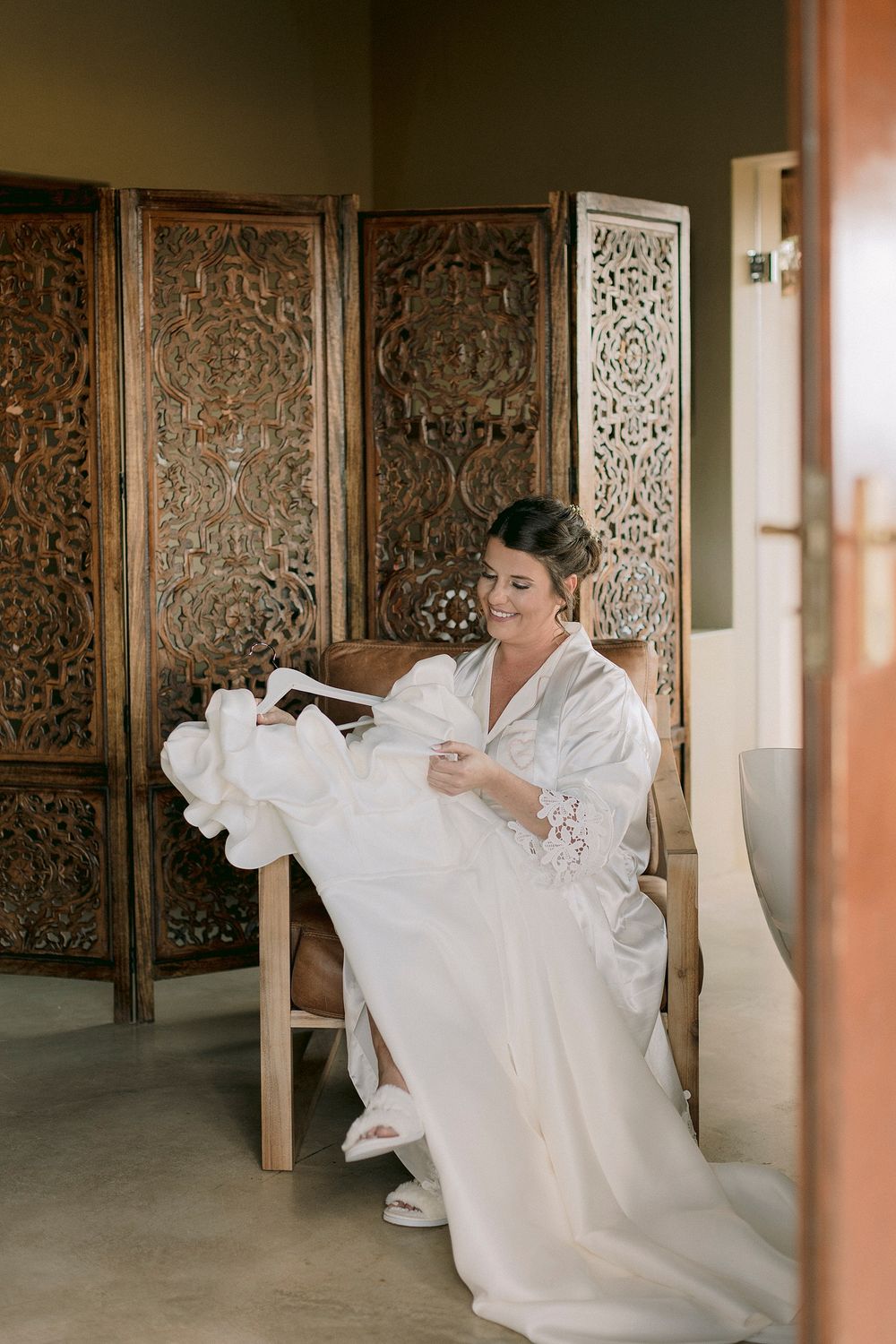Bride in white robe sitting in wooden chair examining her wedding dress before ceremony with ornate screen backdrop.