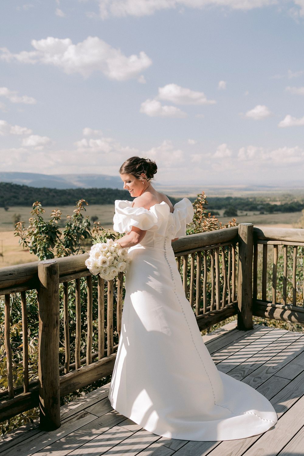 Bride in elegant off-shoulder white gown with train holds bouquet on rustic deck with scenic landscape.