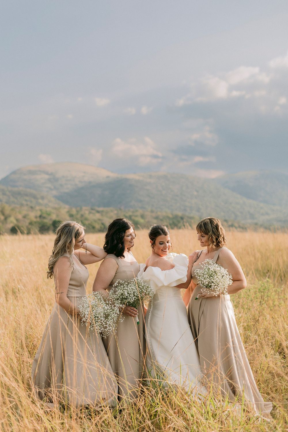 Bride and three bridesmaids in tan gowns holding baby's breath bouquets in golden field with mountain backdrop.