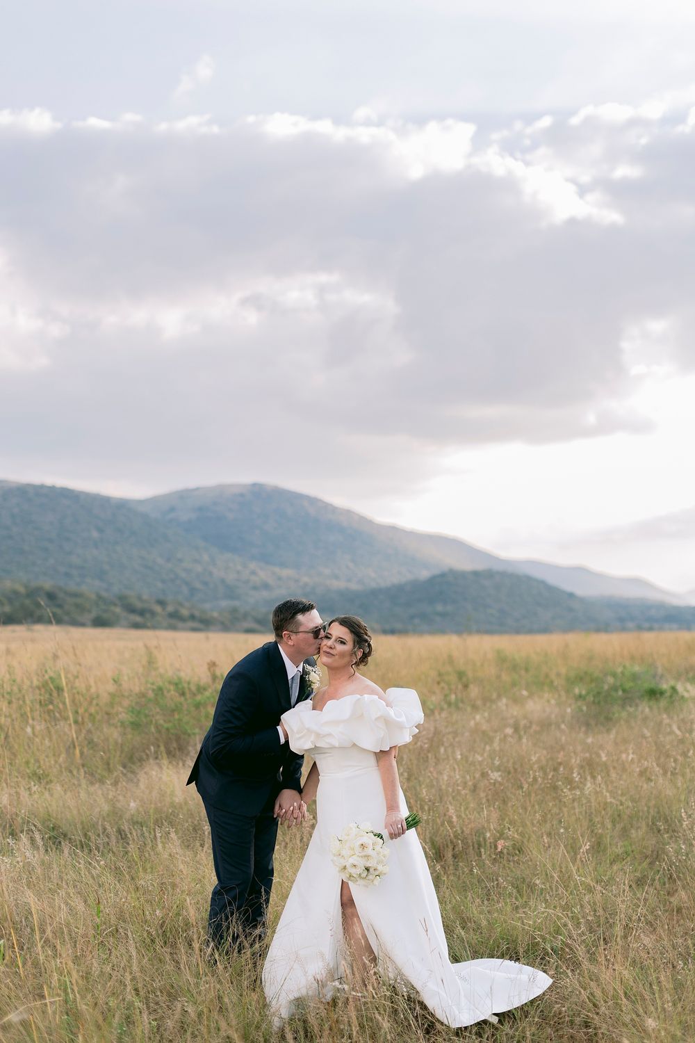 Bride and groom share a kiss in a golden field with mountain backdrop on their wedding day.