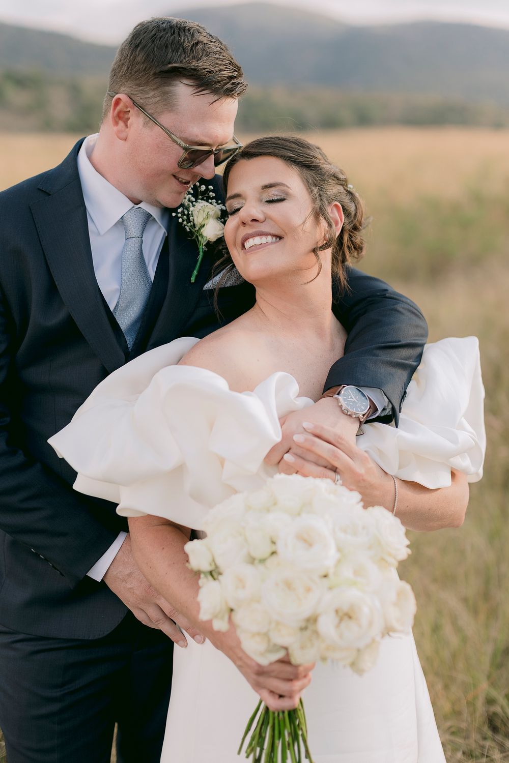 A joyful bride in a white ruffled gown holds a bouquet while her groom in a navy suit embraces her in a golden field.
