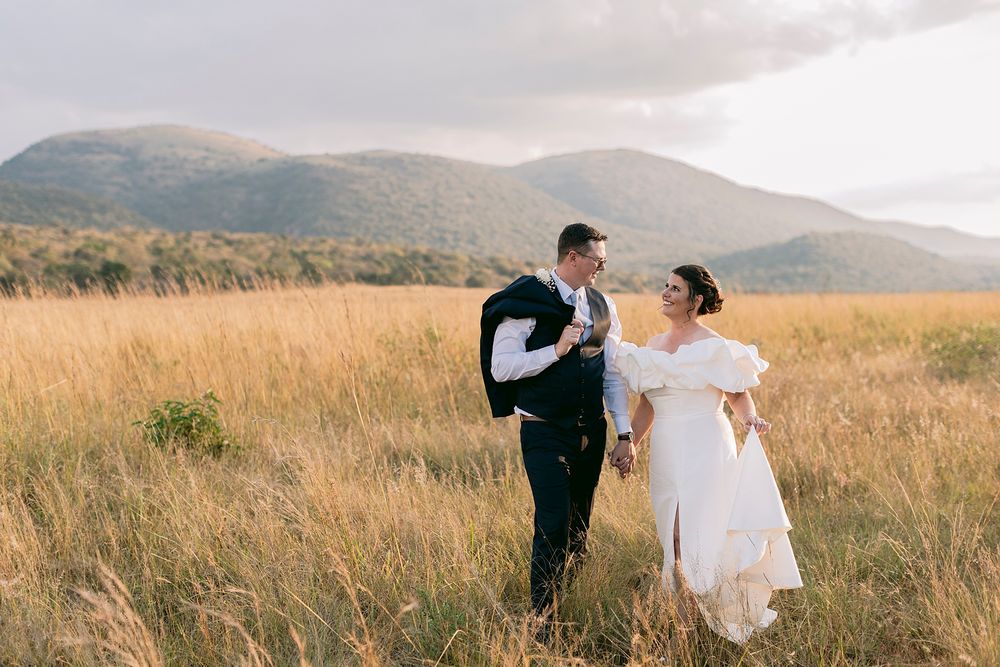 Bride and groom walk hand in hand through golden grass fields with misty mountains backdrop.