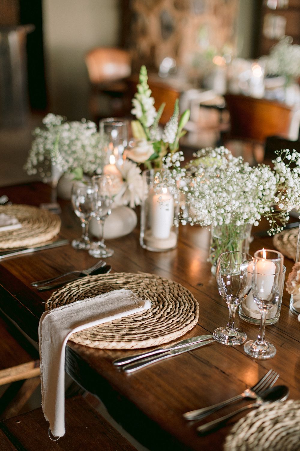 Rustic wedding table setting with woven chargers, baby's breath centerpieces, candles, and crystal glassware on dark wood.