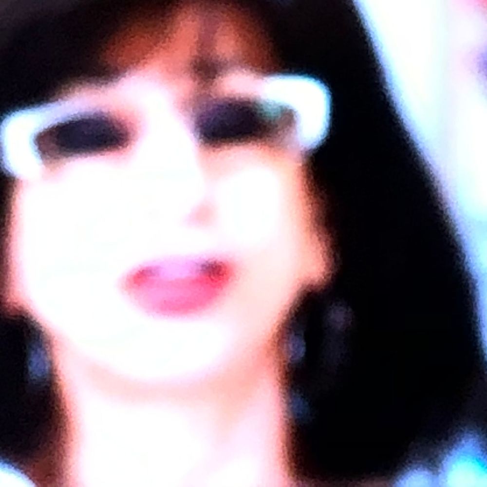 A blurry close-up portrait of a woman with dark hair, white-framed glasses, and red lipstick.