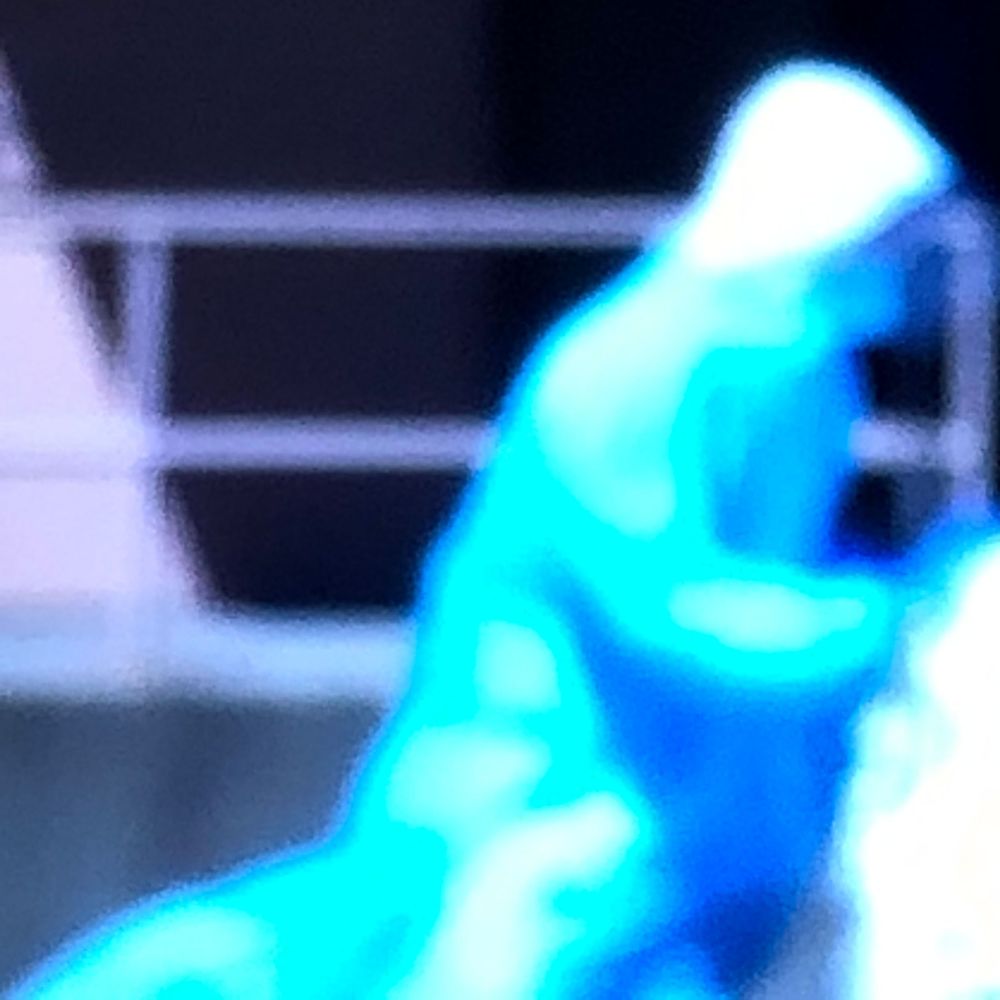 Glowing blue figure in a hooded costume performing on a dark stage with metal railings in the background.