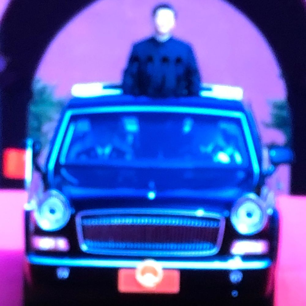 Glowing blue toy limousine with a figure standing through the sunroof, illuminated against a purple background.