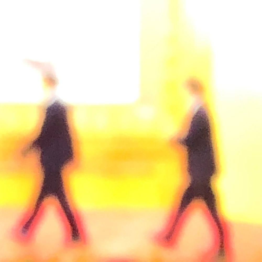 Two blurred figures in dark suits with red shoes walking against a bright yellow background.