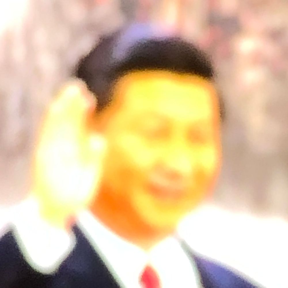 A man in a dark suit with a red tie waves and smiles in a blurred outdoor setting.