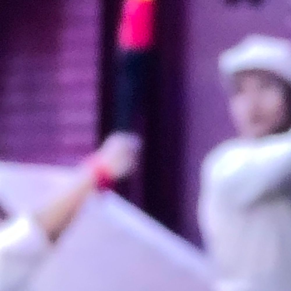 Blurry image of a person in white outfit holding a glowing pink light stick on a purple stage.