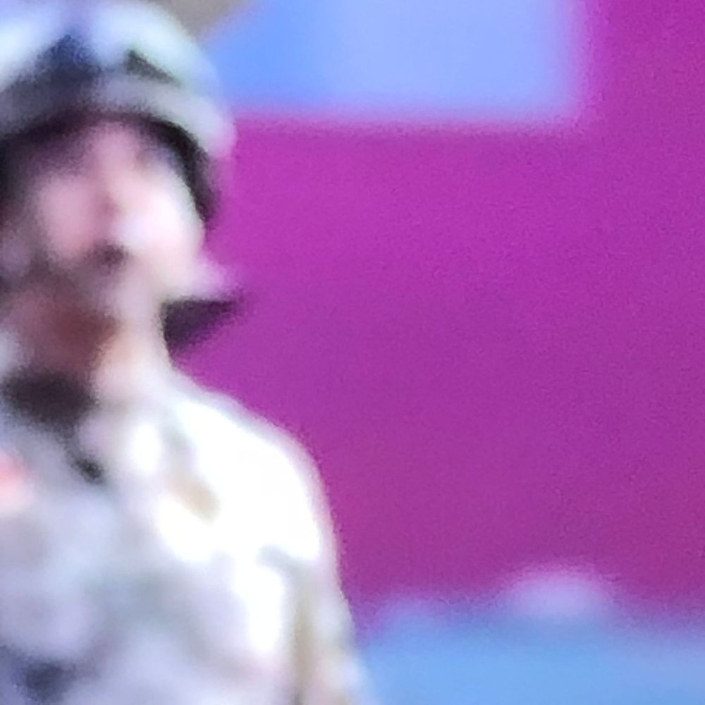 Blurry image of a soldier in camouflage uniform and helmet standing against a bright pink and blue background.