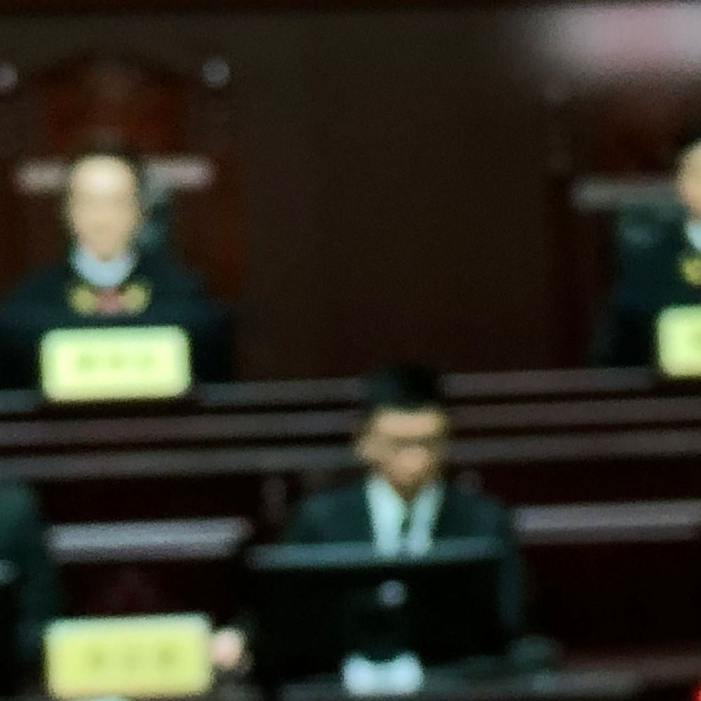 Blurry courtroom scene with defendants wearing yellow placards seated before a judge in a formal setting.