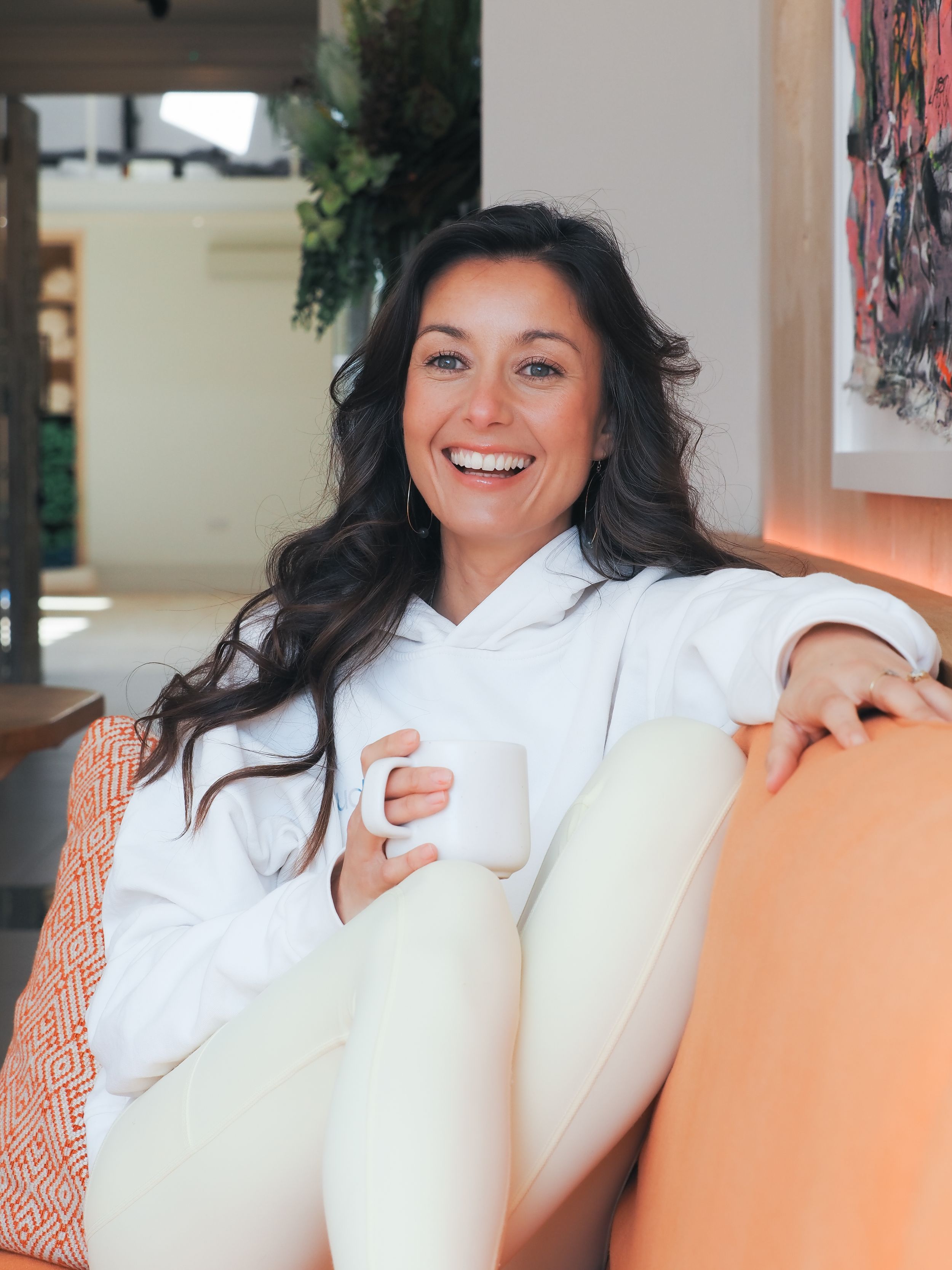 Person relaxing on couch in white hoodie with coffee mug and warm smile in bright living room setting.