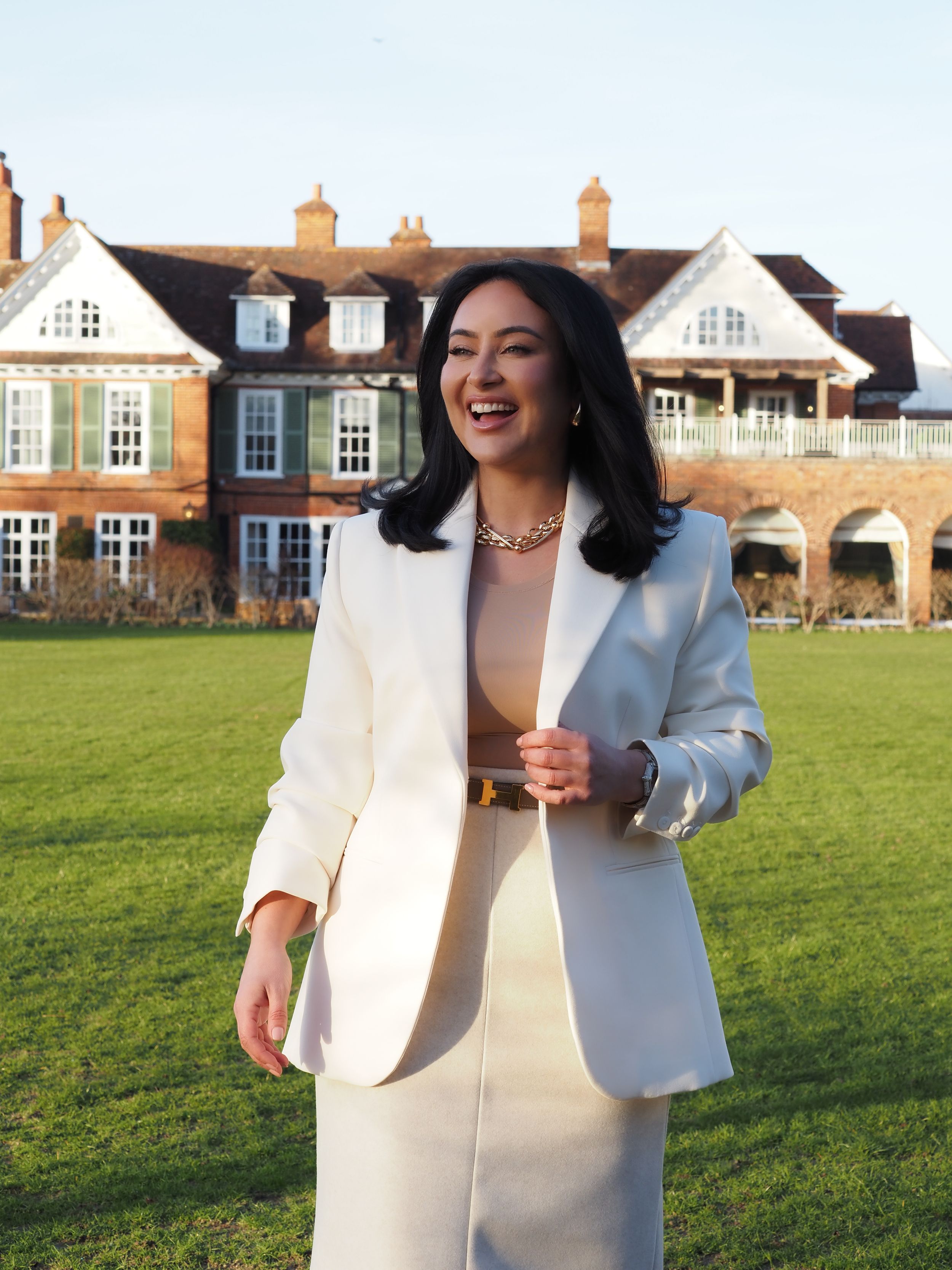 A person in a cream-colored business suit stands smiling on a grass lawn in front of a grand brick mansion.