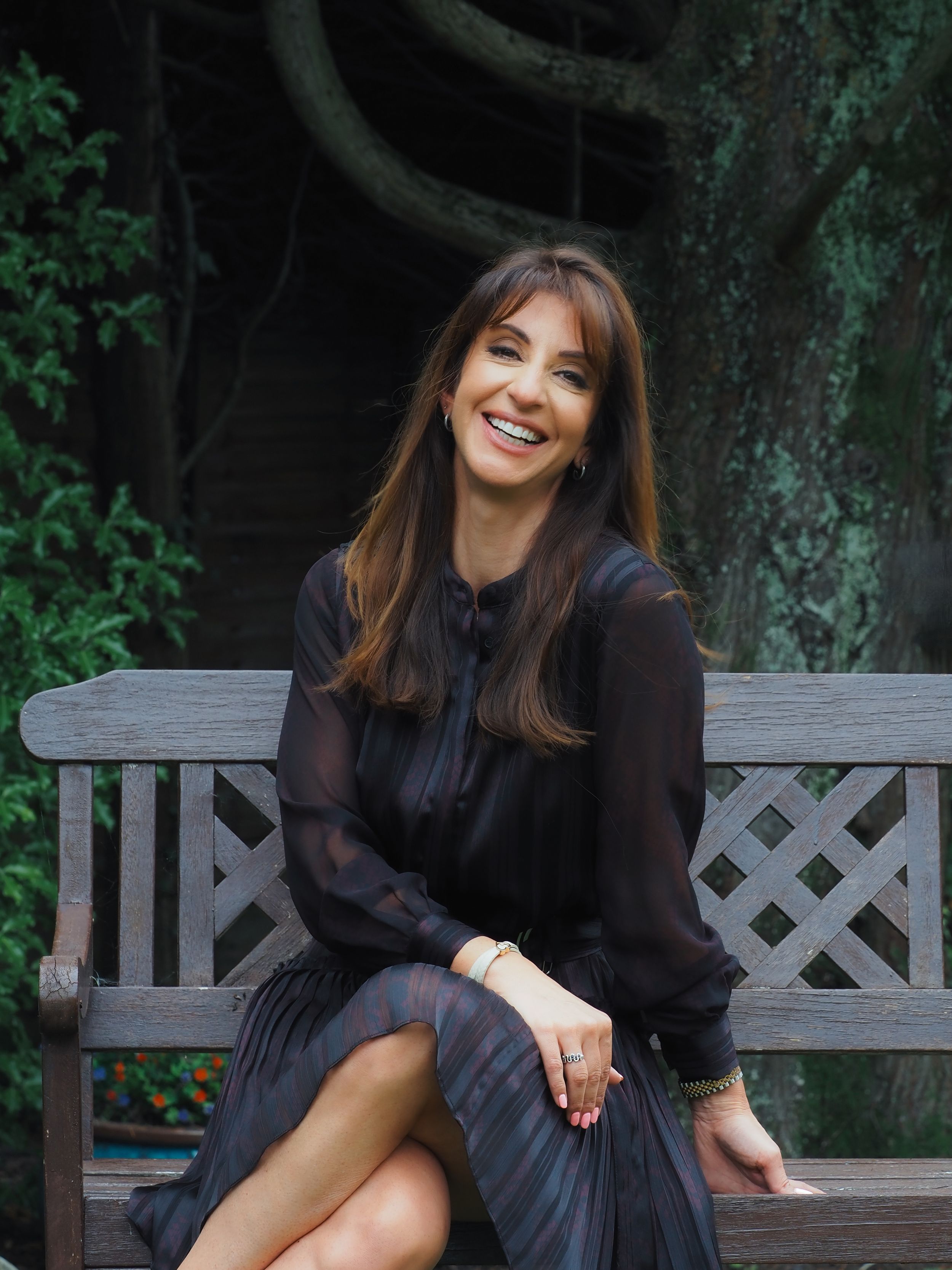 A person in a black flowing dress sits smiling on a wooden garden bench surrounded by lush greenery.